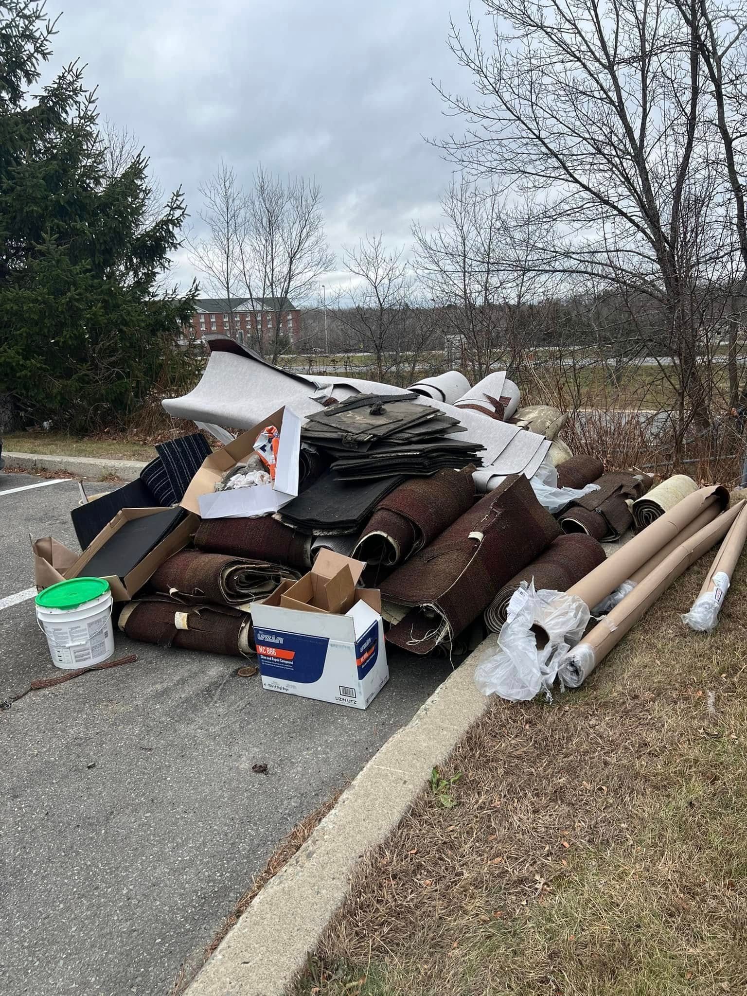 Piles of roofing debris and trash dumped along a roadside shoulder near trees and a field.