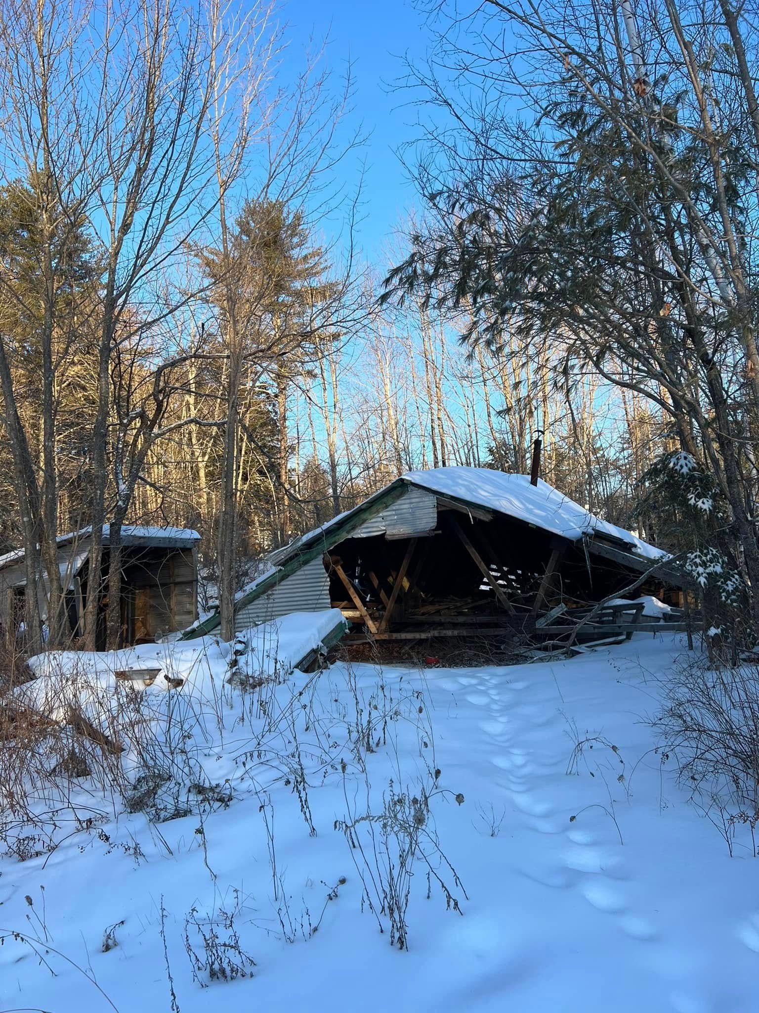 Snow-covered cabin partially collapsed in a wooded winter landscape under a clear blue sky