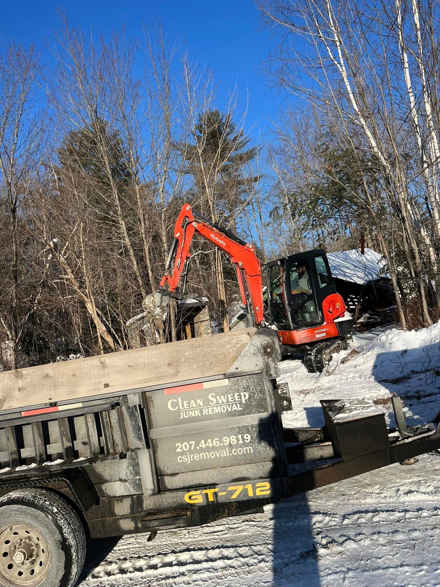 Orange excavator loading dirt into a dump truck in a snowy wooded worksite