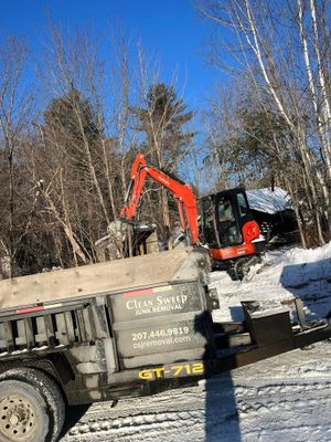 Excavator loading a dump truck in a snowy wooded area under a clear blue sky