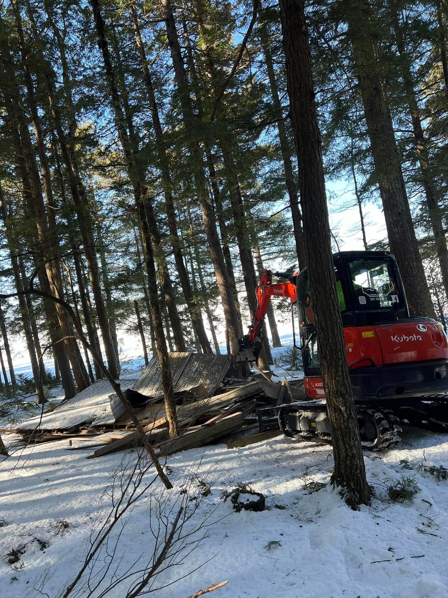 Excavator clearing snow among tall trees in a wooded area