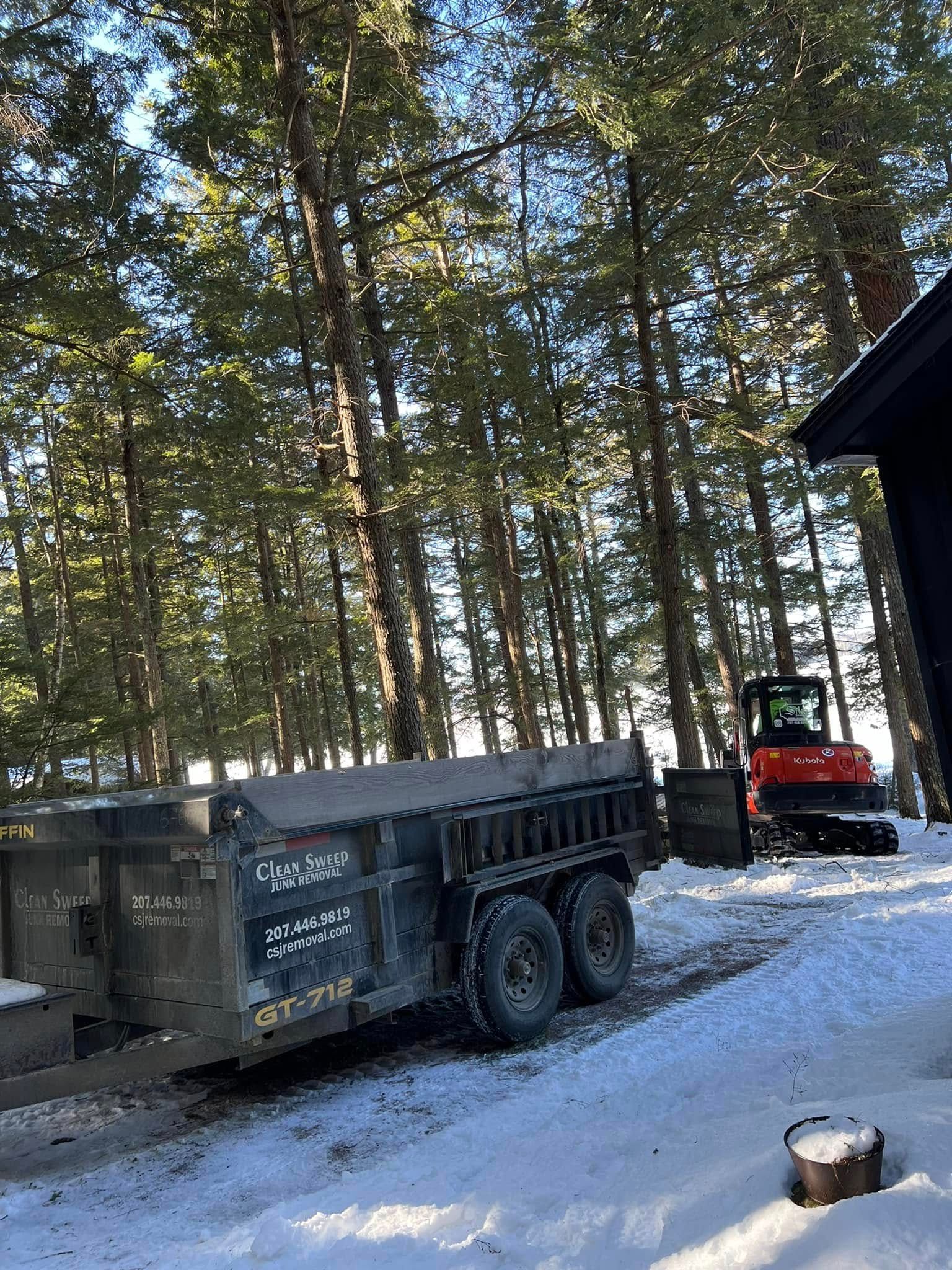 Dump trailer and red tractor in snowy wooded yard beside a dark building.