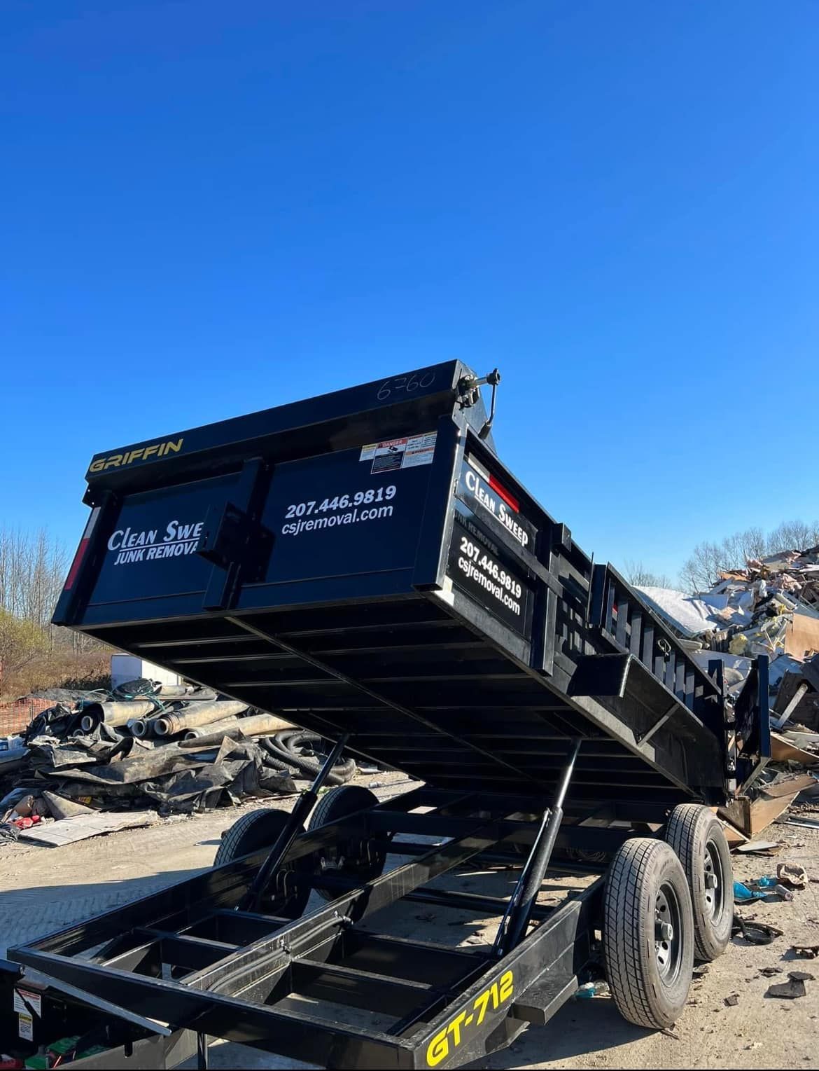 Black dump trailer tipped steeply upward in a rubble-strewn construction debris site under a clear blue sky