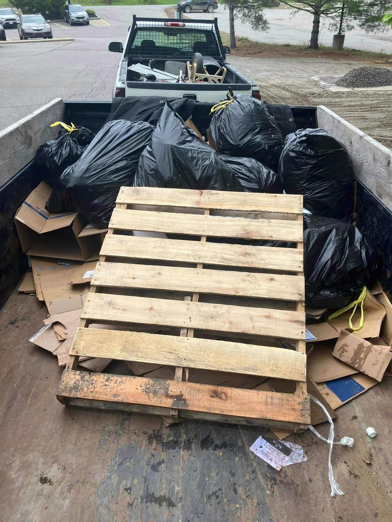 Pickup truck bed filled with black trash bags, cardboard, and a wooden pallet at a curbside area