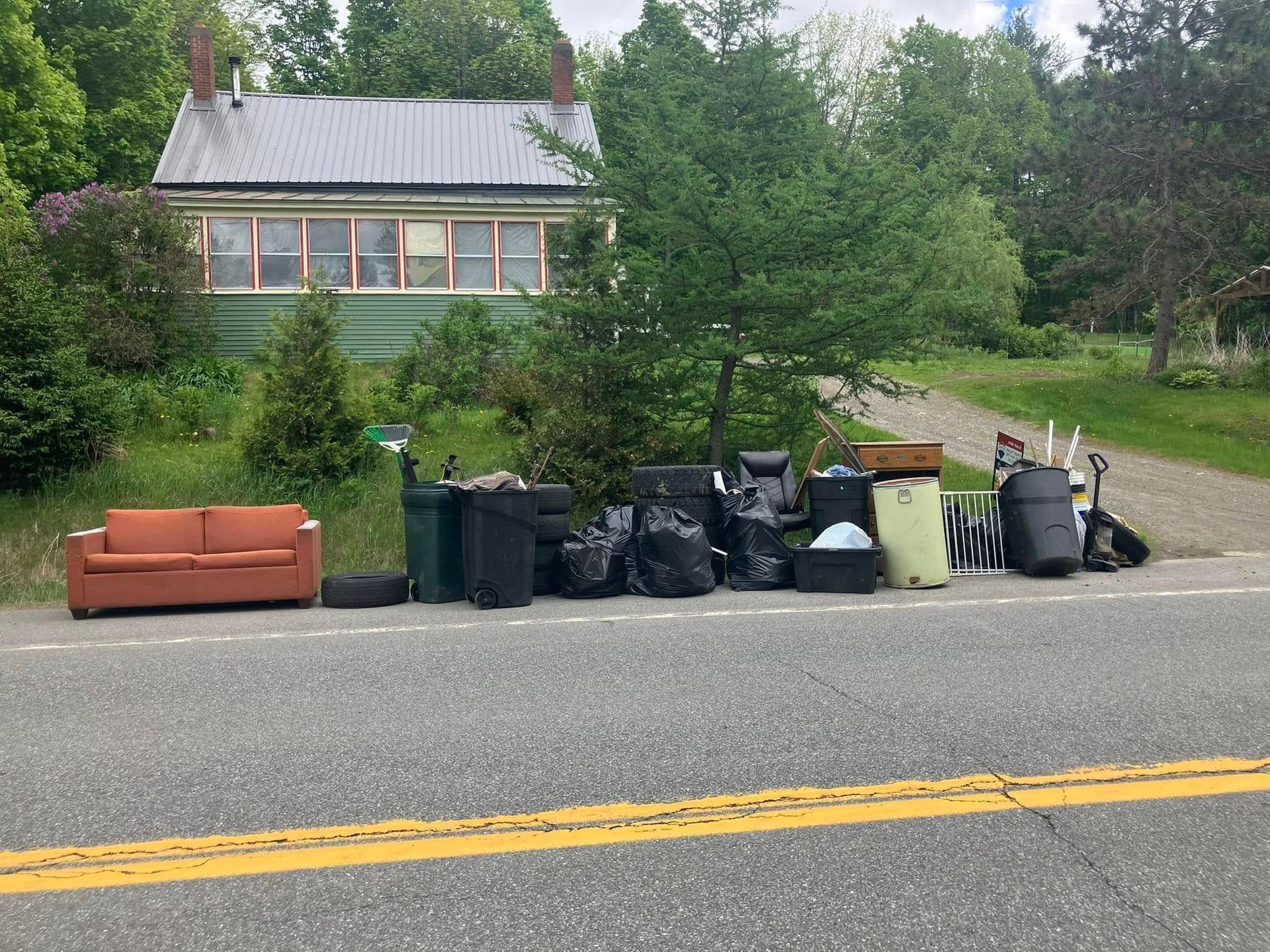 Curbside trash and recycling bins with an orange couch on a roadside in front of a house and trees.