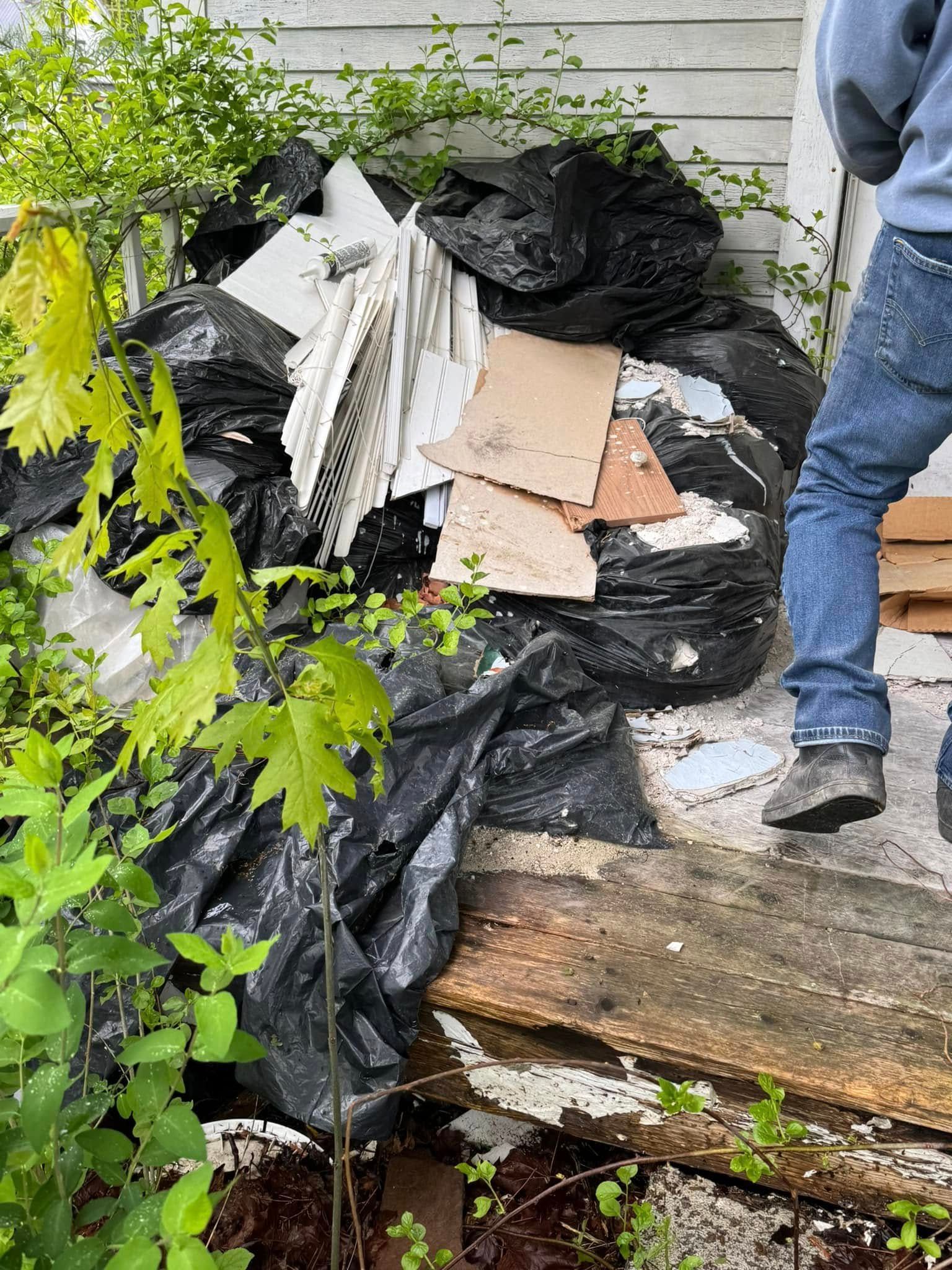 Pile of trash bags, cardboard, and broken white slats beside a house, with a person standing nearby.