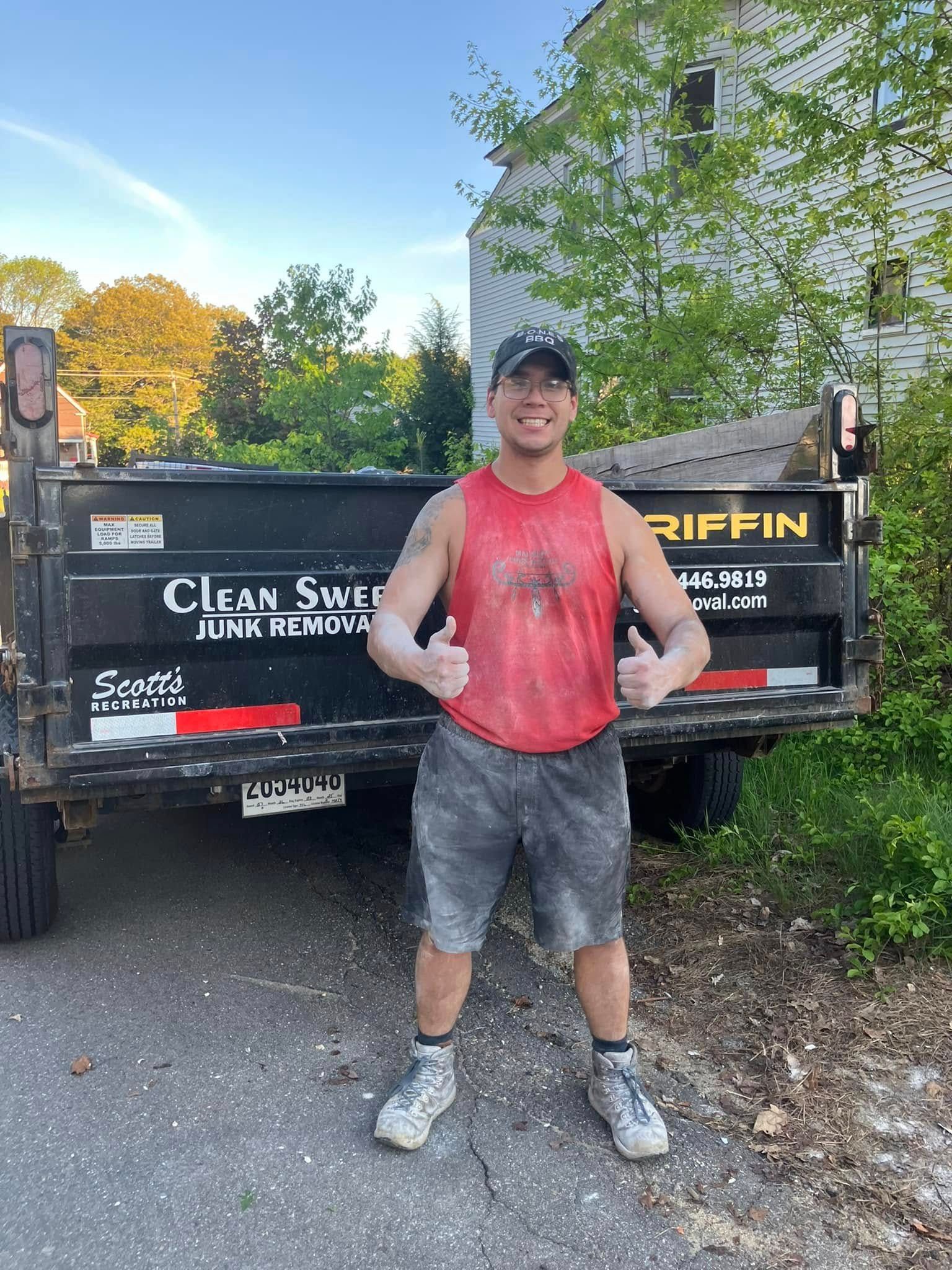 Man standing in a red shirt and cap in front of a dump trailer outdoors.