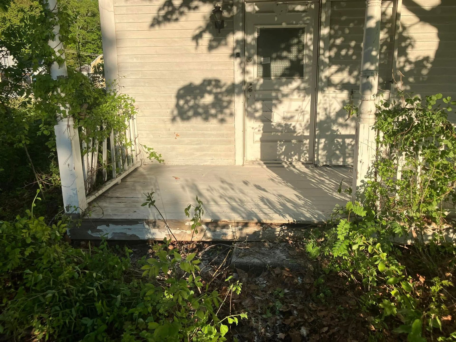 Small porch with wooden steps and white railing beside a beige house, surrounded by greenery.