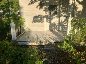 Small wooden porch with steps, white railing, and a beige house surrounded by dense greenery.