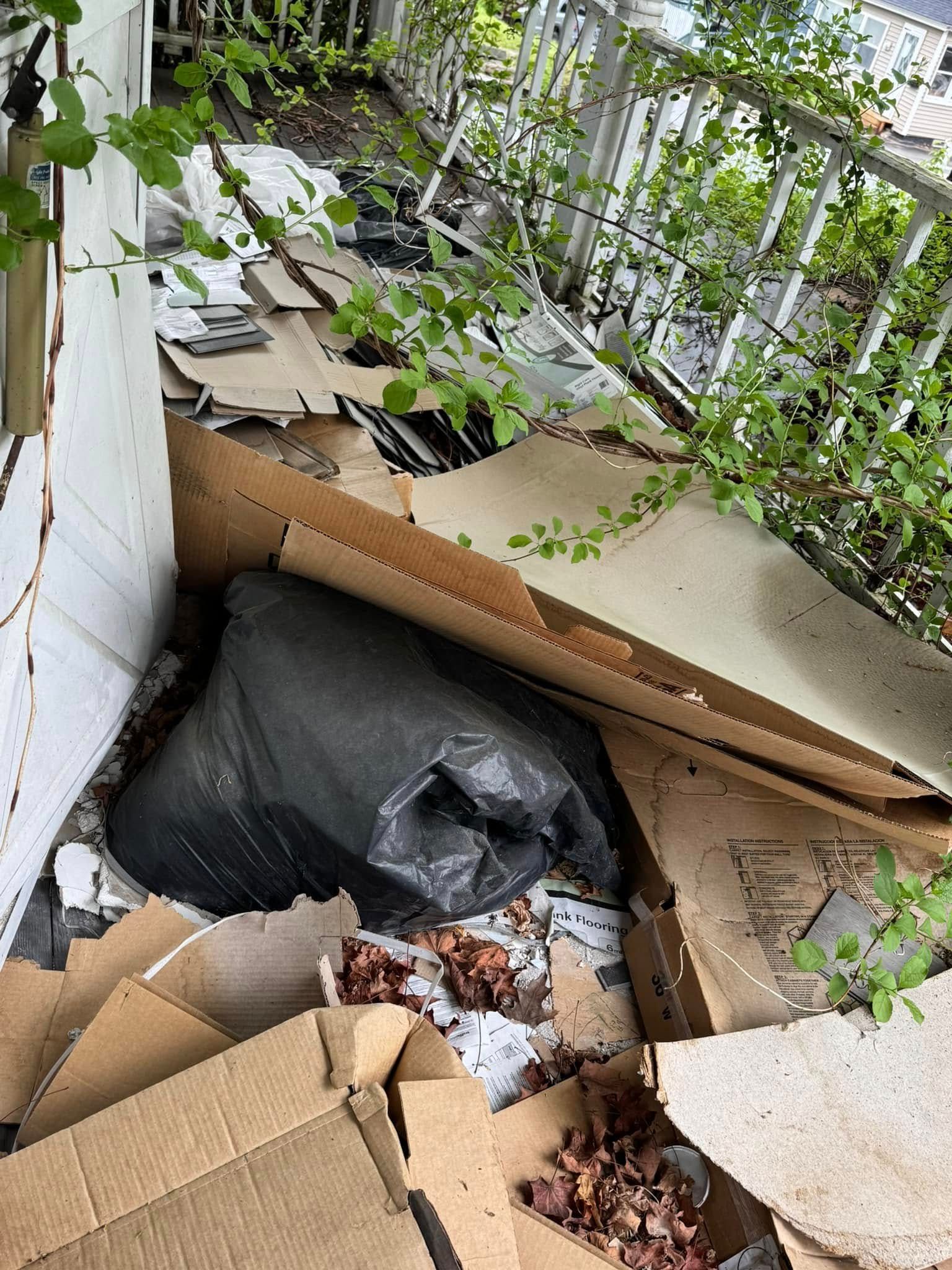 Cardboard boxes and black trash bags clutter a narrow outdoor walkway beside a white wall and green plants.