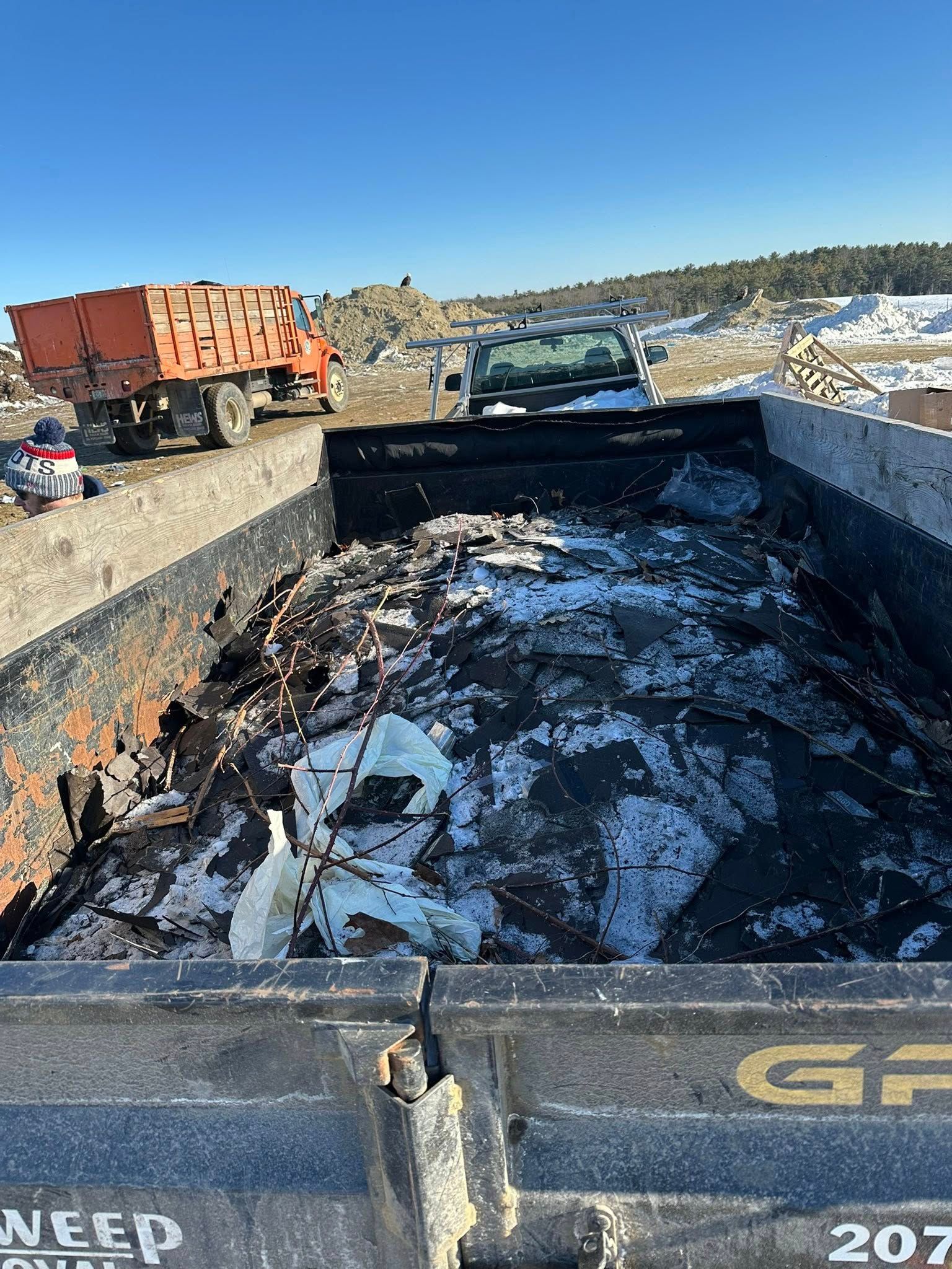 Burned debris in a dump truck bed at a demolition site, with a truck and snowy hills in the background