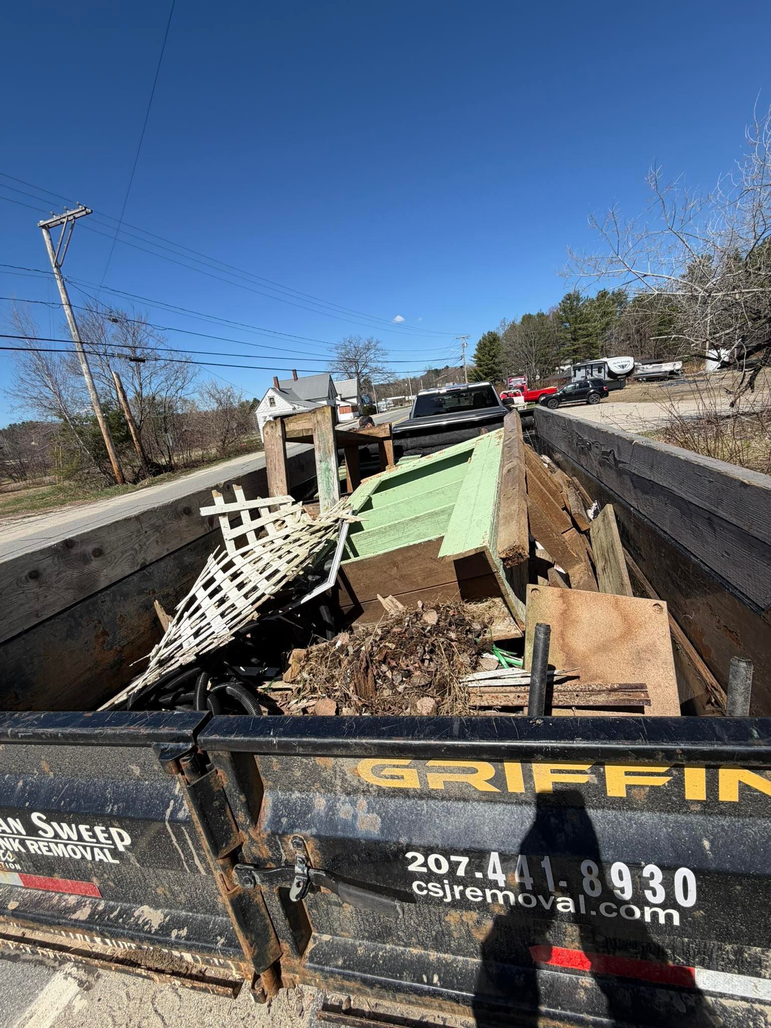 Dumpster of construction debris on a road, with a green plank and black dumpsters under a clear blue sky