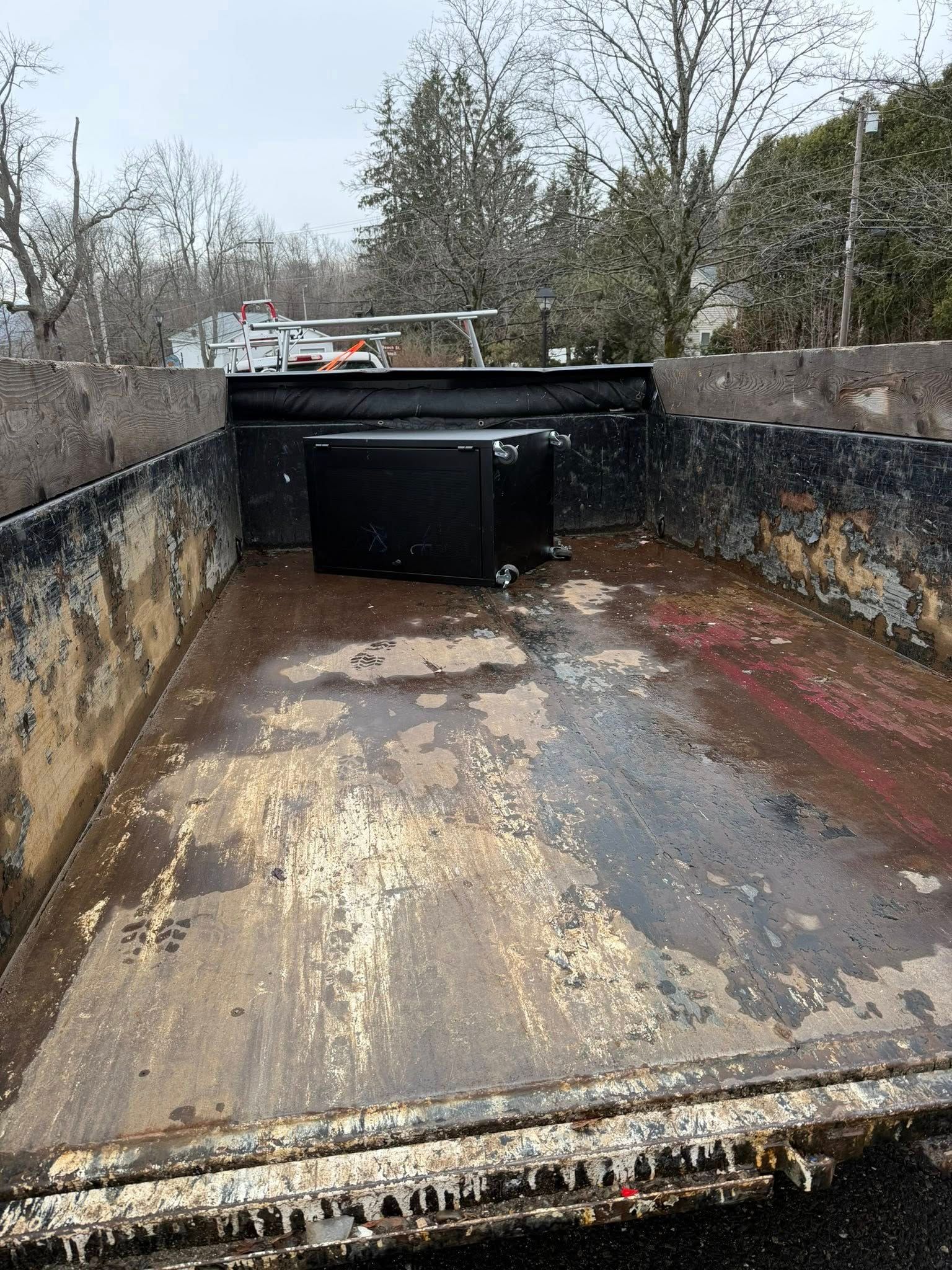 Rusty empty dump truck bed with black tailgate, seen outdoors behind bare trees.
