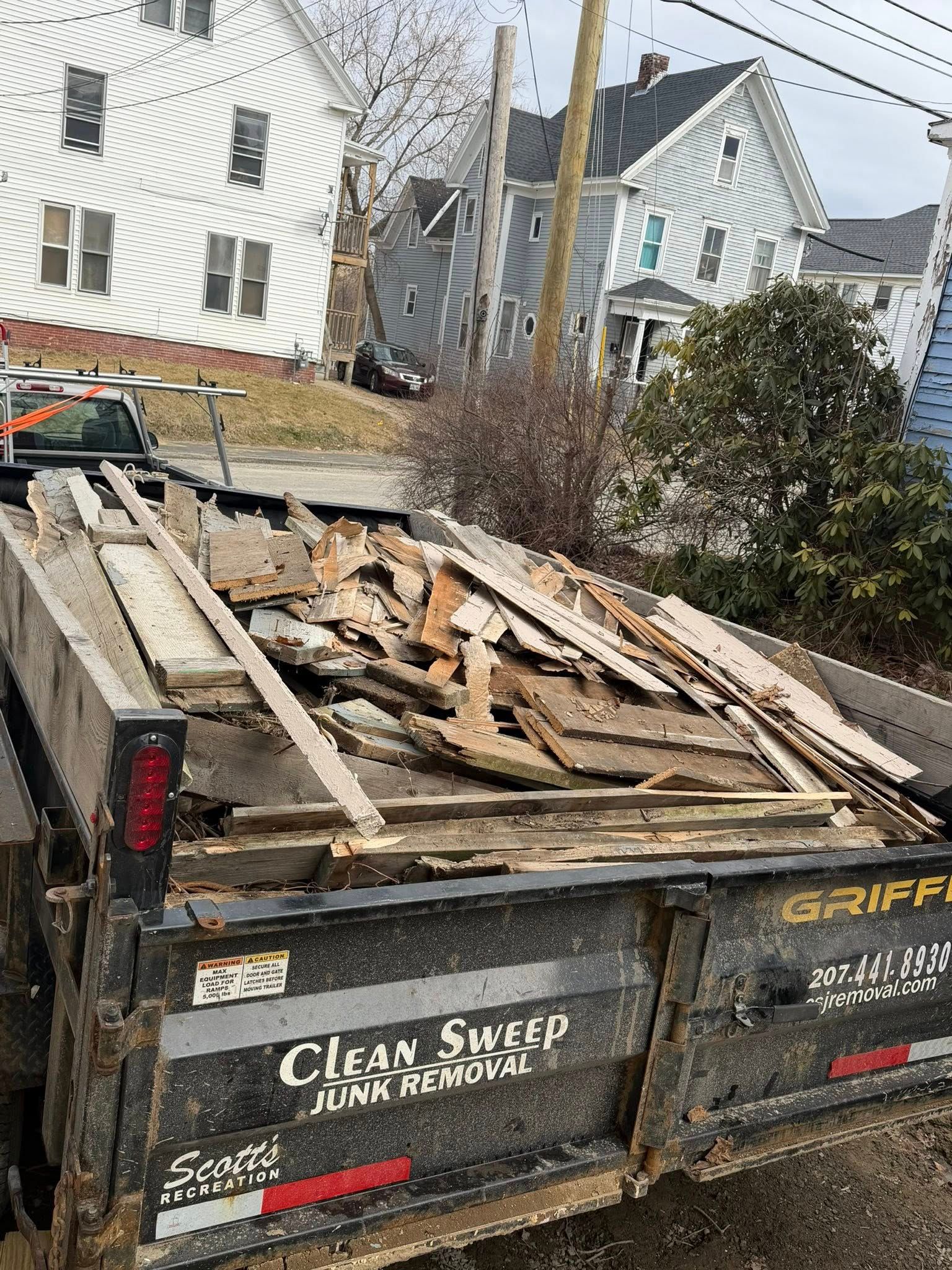 Dump trailer loaded with broken wood debris beside damaged white houses and utility poles