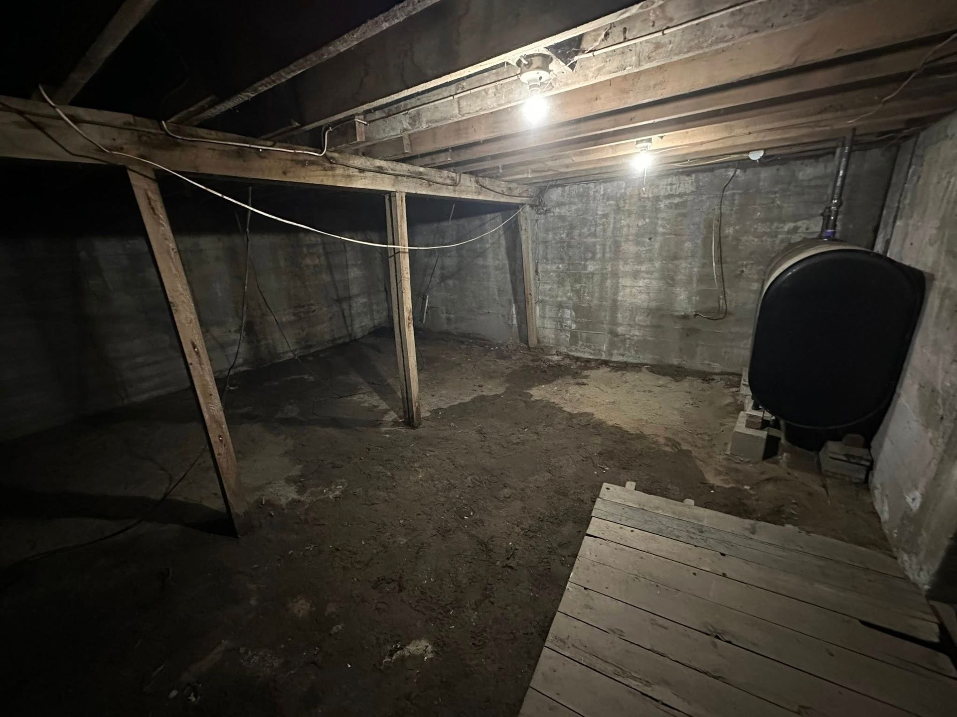 Dim unfinished basement with exposed beams, dirt floor, and a black tank or barrel against the wall.
