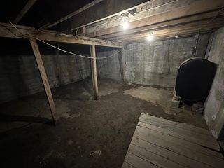 Dim unfinished basement with exposed beams, dirt floor, and a black tank or barrel against the wall.
