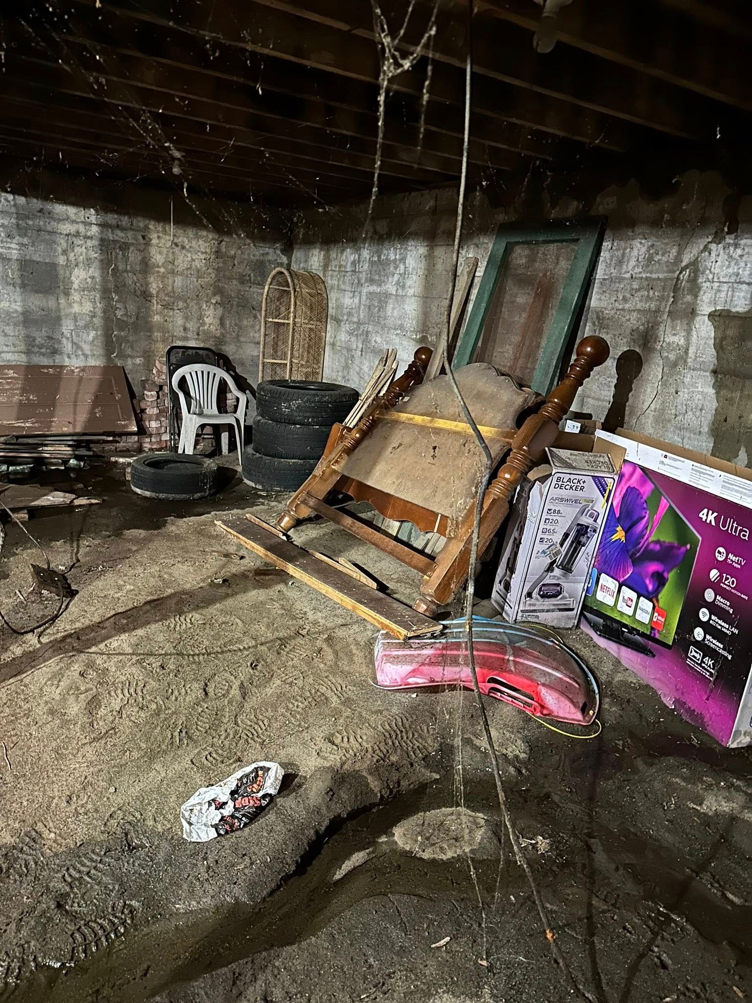 Flooded basement with debris, a tipped wooden chair, stacked tires, and a colorful board leaning against the wall.