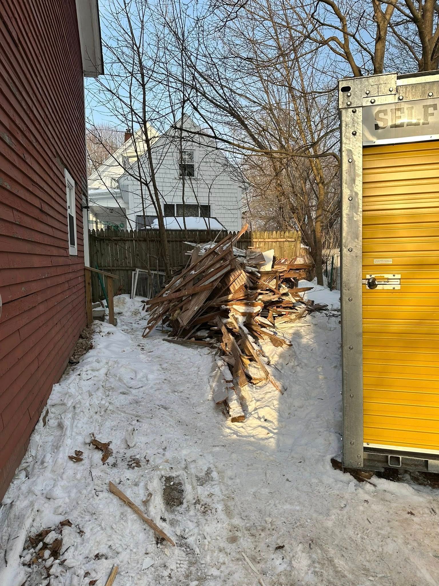 Snowy alley beside red brick wall and yellow storage container, with piled branches and a house in the background