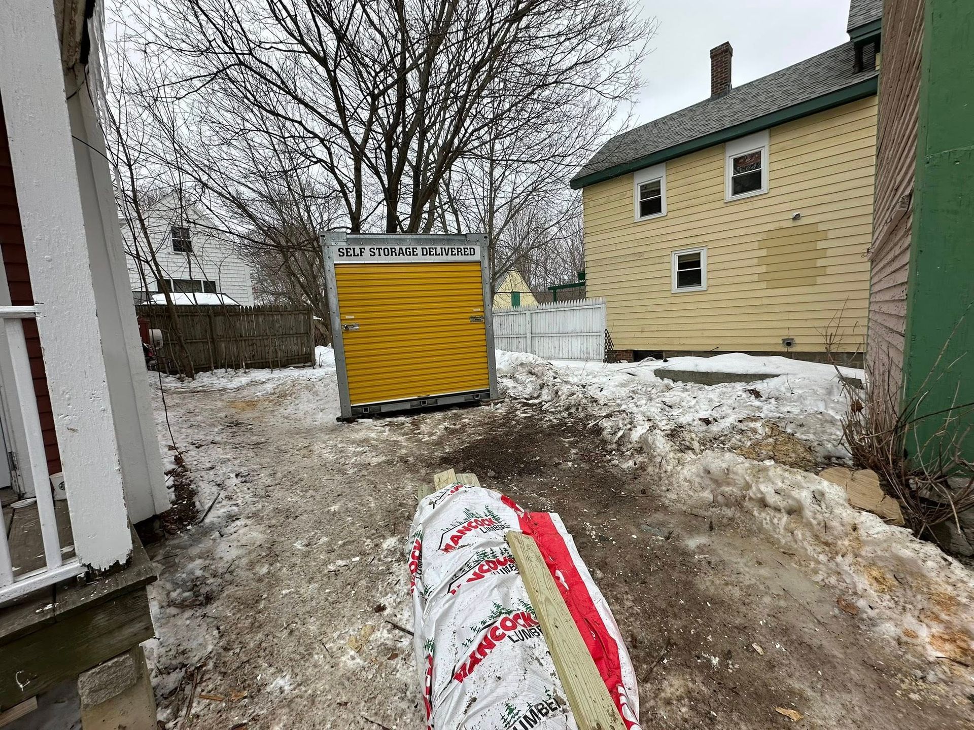 Snowy backyard with a yellow shed, a house, and a bag of materials in the foreground