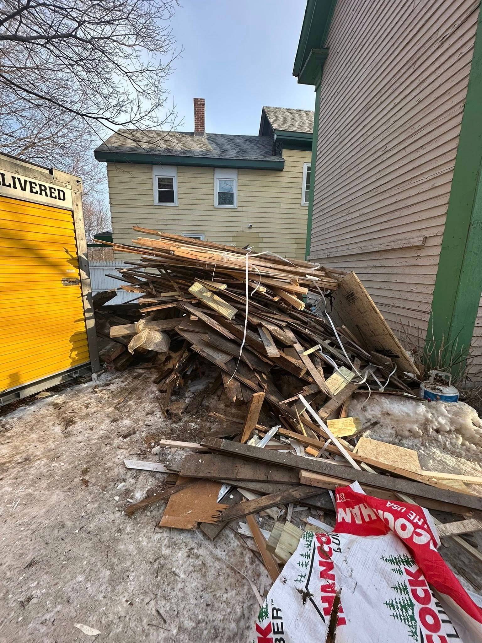 Pile of construction debris beside a house, with a yellow dumpster on the left and siding on the right.