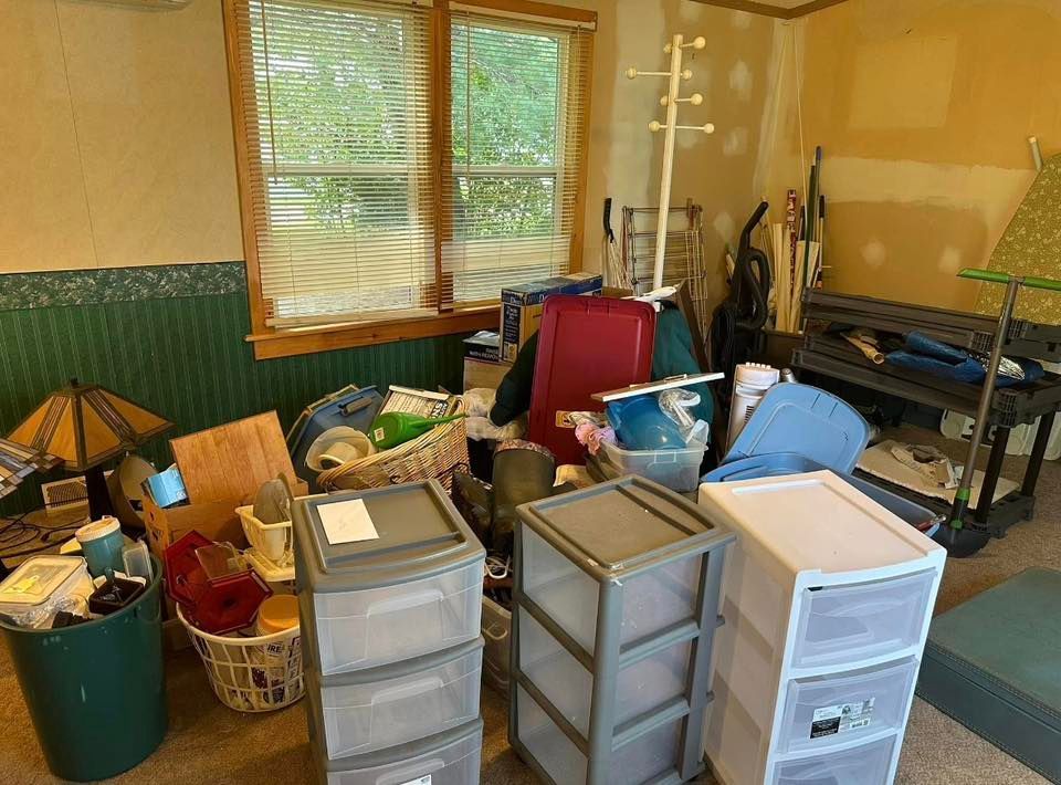 Cluttered room with stacked plastic drawers, boxes, tools, and bags beside a window with blinds