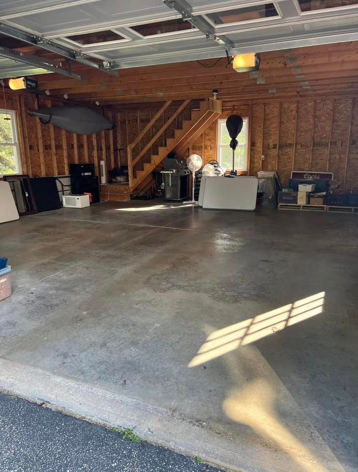Empty garage with concrete floor, wooden stairs, and stored items along the back wall.