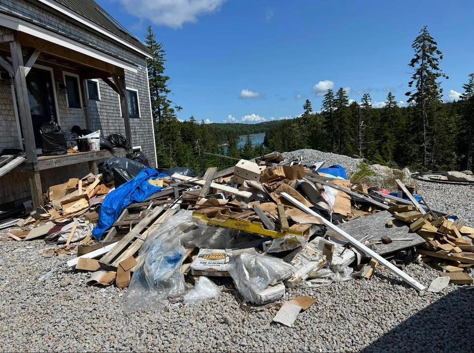 Pile of construction debris beside a house with blue tarp, gravel driveway, and trees in the background