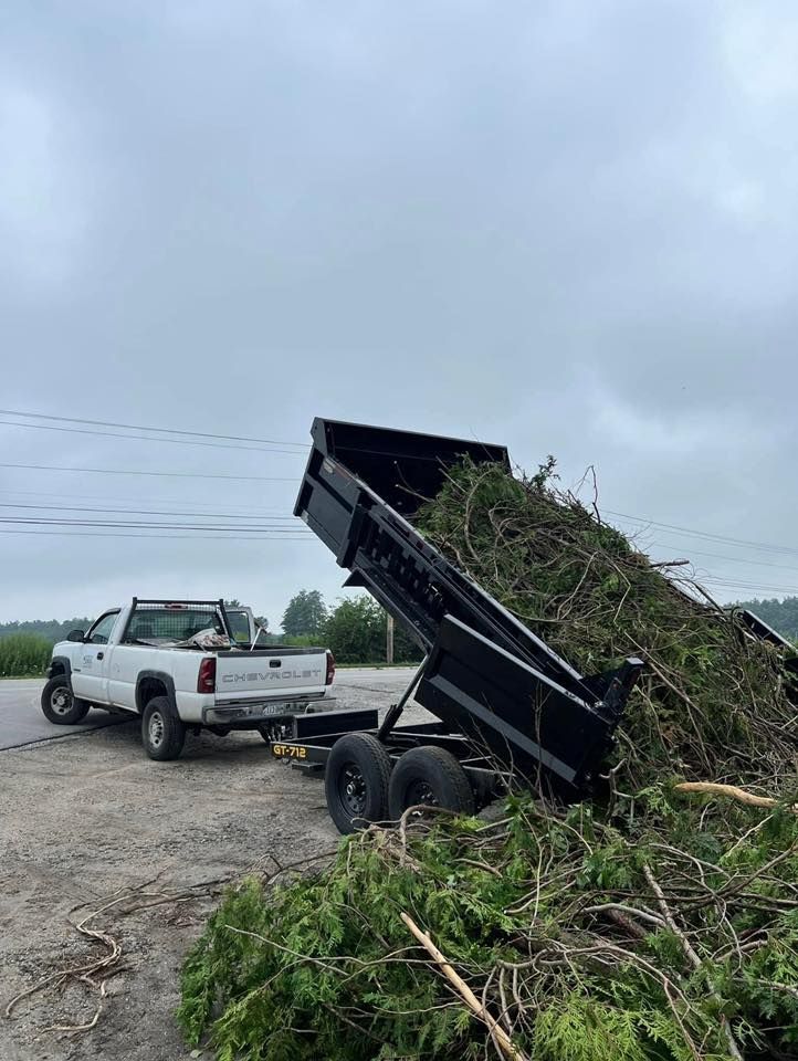 Pickup truck unloading brush into a dump trailer on a gravel roadside under cloudy skies