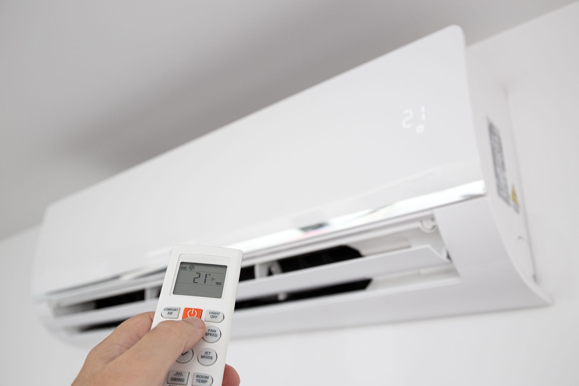 A hand operates a remote control of a domestic air conditioning unit mounted on a wall.