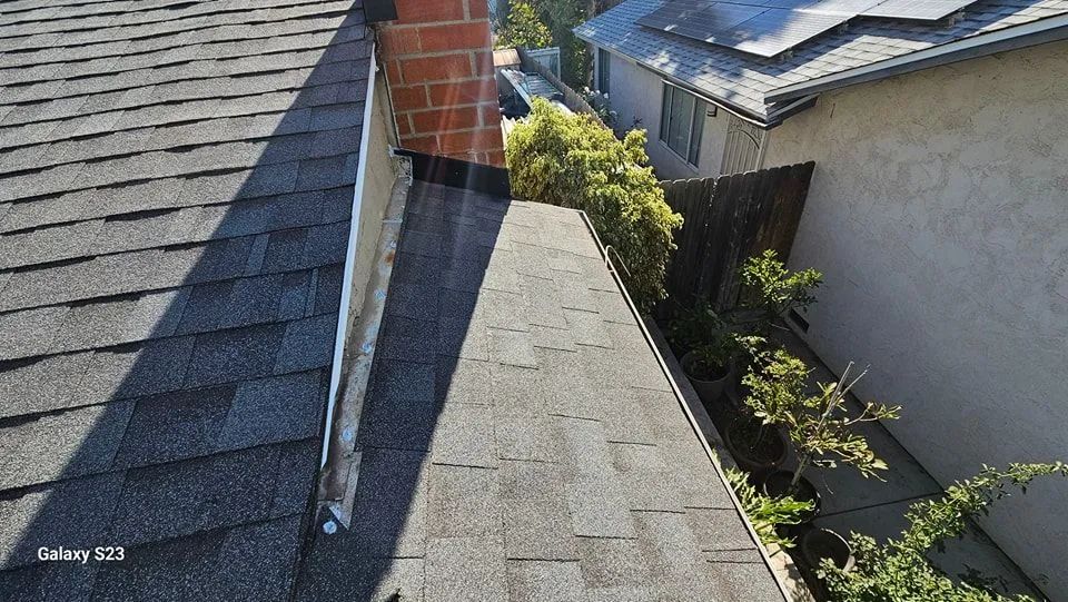 Rooftop with asphalt shingles and a brick chimney, viewed along a narrow sloped roof between houses.