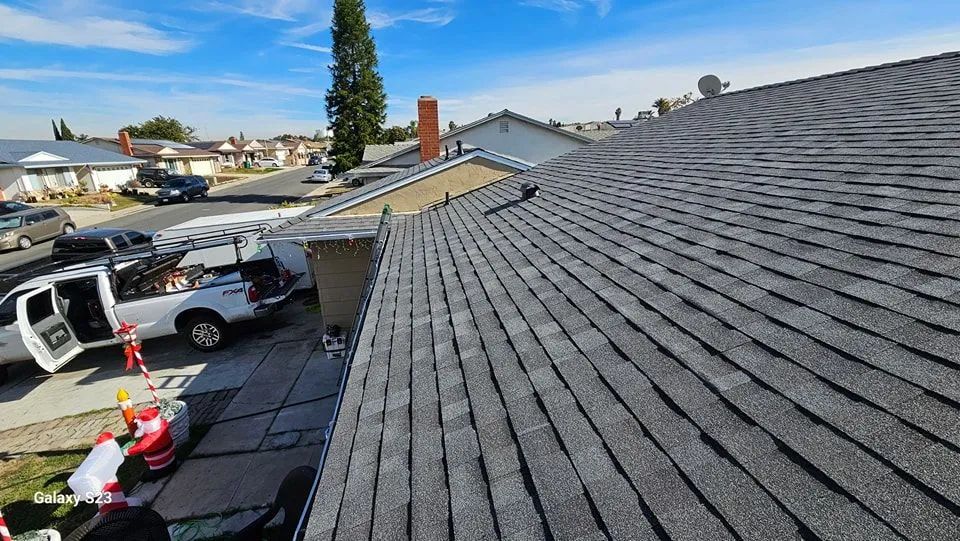 Roofline view of a suburban street with asphalt shingles, parked cars, and a worker in a safety harness