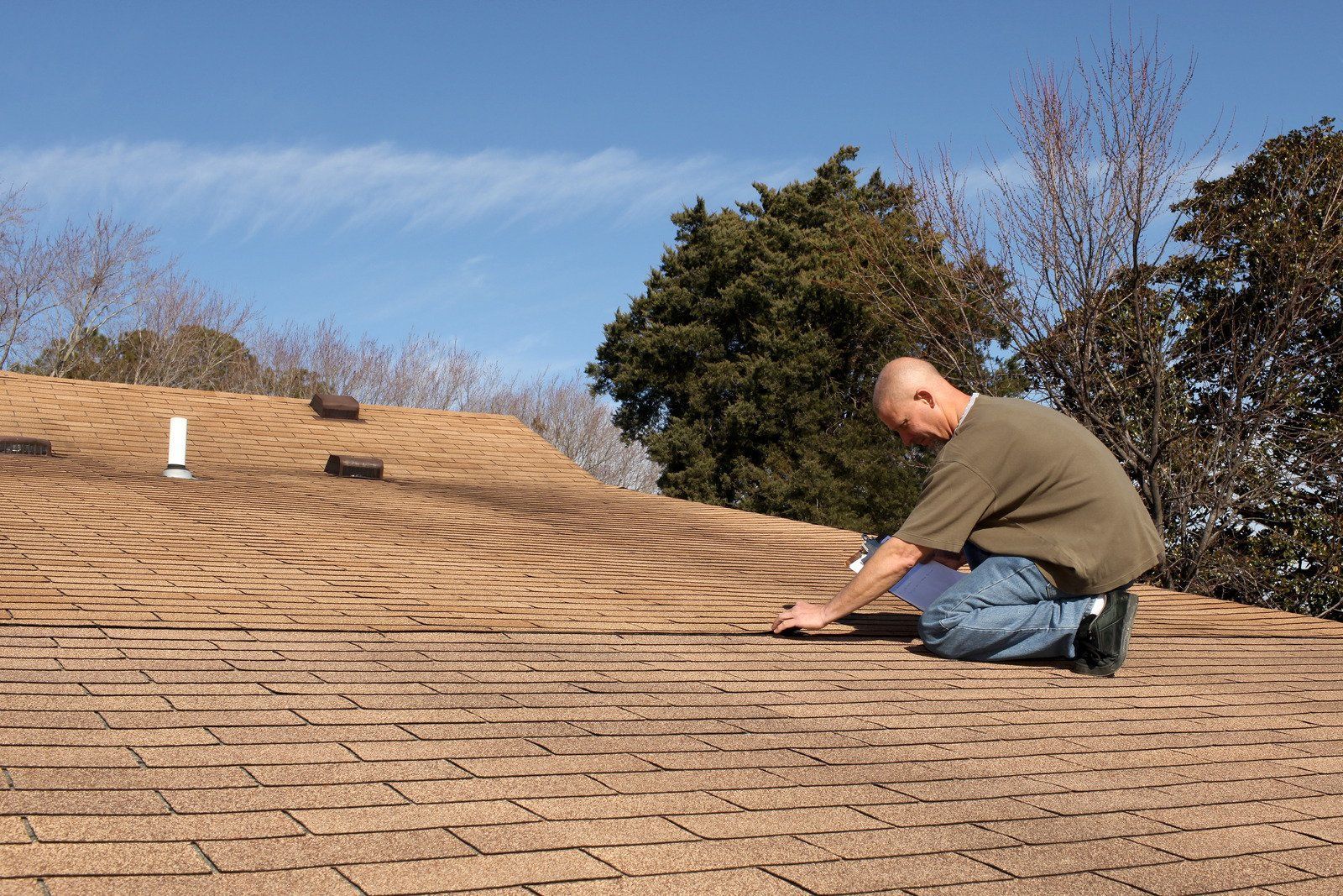 Man inspecting a brown shingle roof on a sunny day; trees and blue sky in the background.