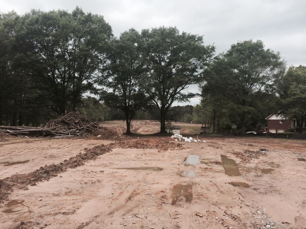 Cleared dirt lot with puddles, trees in background, pile of logs on the left, and building on right.