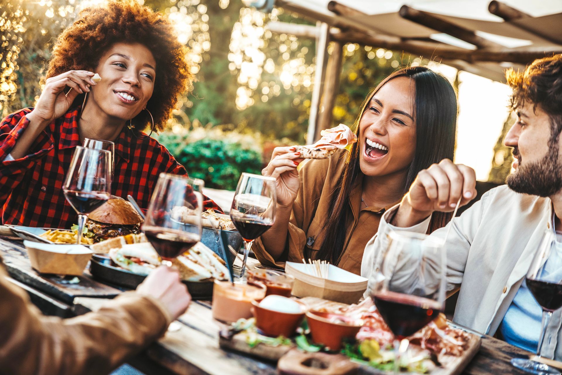 Friends enjoying food and wine outdoors, laughing together at a wooden table. Sunlight.