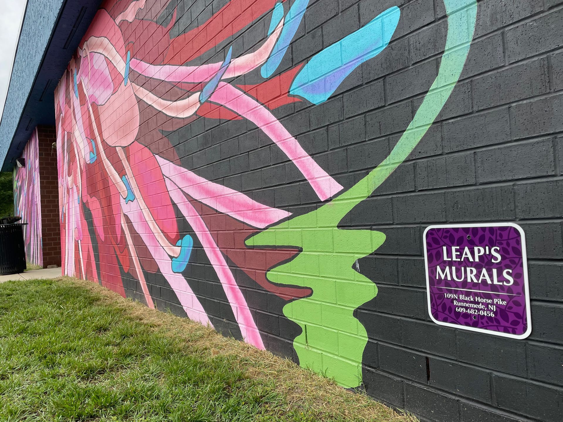 Colorful abstract mural on a gray brick wall with a small sign reading “Leapi’s Murals” near grass.