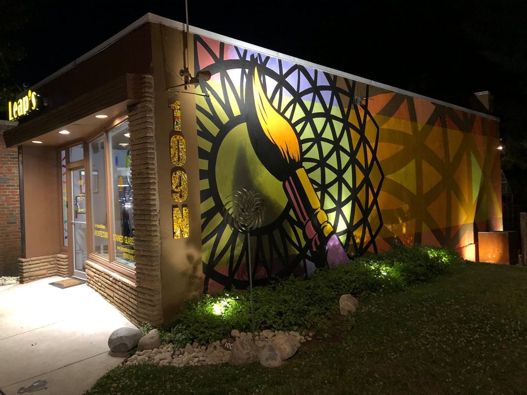 Nighttime view of a storefront with a large, colorful mural of a paintbrush against a geometric, stained-glass background.