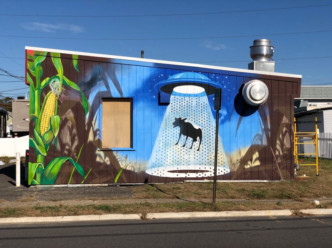 A colorful mural on a small building depicts a UFO beaming up a cow from a cornfield against a bright blue sky.