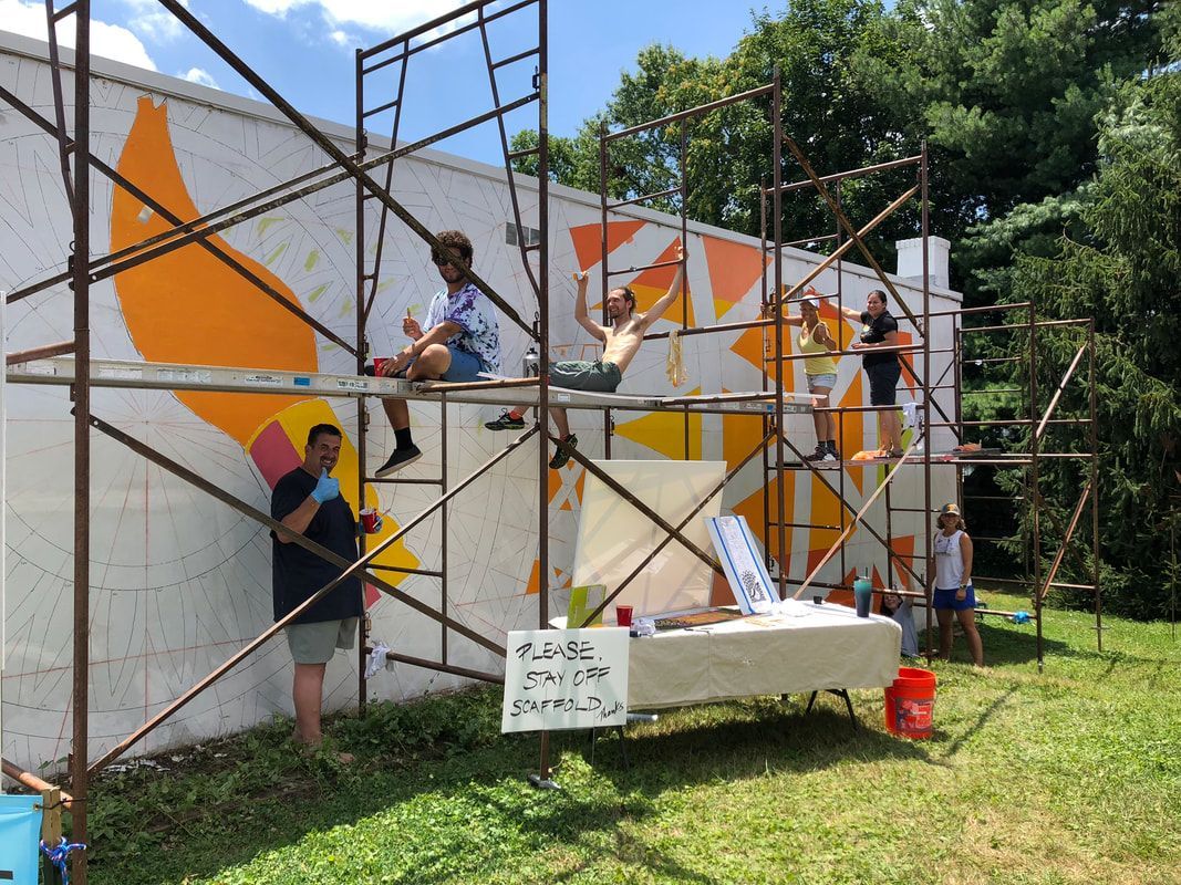 A group of people painting a large outdoor mural on a white wall using yellow and orange paint from scaffolding.