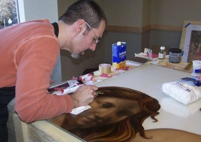 A person in an orange long-sleeved shirt carefully paints a detailed portrait of a face onto a surface on a table.