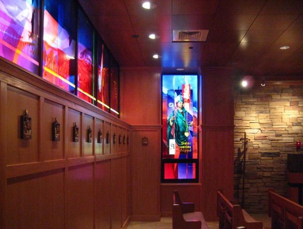 Interior of a chapel with wood-paneled walls, vibrant stained-glass windows, and rows of pews facing a stone accent wall.