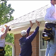 Three workers lifting a white roof panel onto a house under construction.