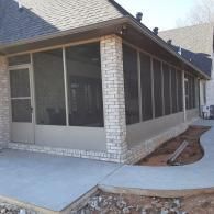 Corner view of a screened porch with stone columns, concrete patio, and adjacent walkway beside a house
