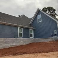Blue-sided house with stone foundation and red dirt landscaping under an overcast sky