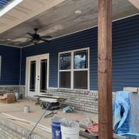 Covered porch with blue siding, brick base, fan, windows, and construction materials on the floor.