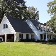 White house with dark roof and dormer windows on a sunny grassy lawn beside trees