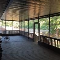 Covered wooden porch with railing and ceiling fan, overlooking trees through screened openings.