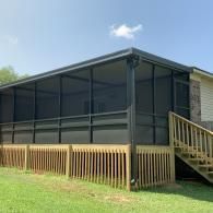 Screened-in porch attached to a house with wooden stairs and a fenced yard under a blue sky