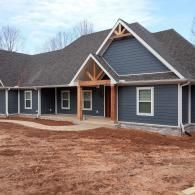 New blue-gray house with white trim and front porch, set on a bare dirt lot under a cloudy sky