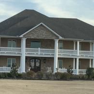 Large two-story house with white columns and balcony, dark roof, and stone facade behind a driveway