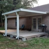 Back patio with a covered wooden deck attached to a brick house, featuring chairs and a grill.